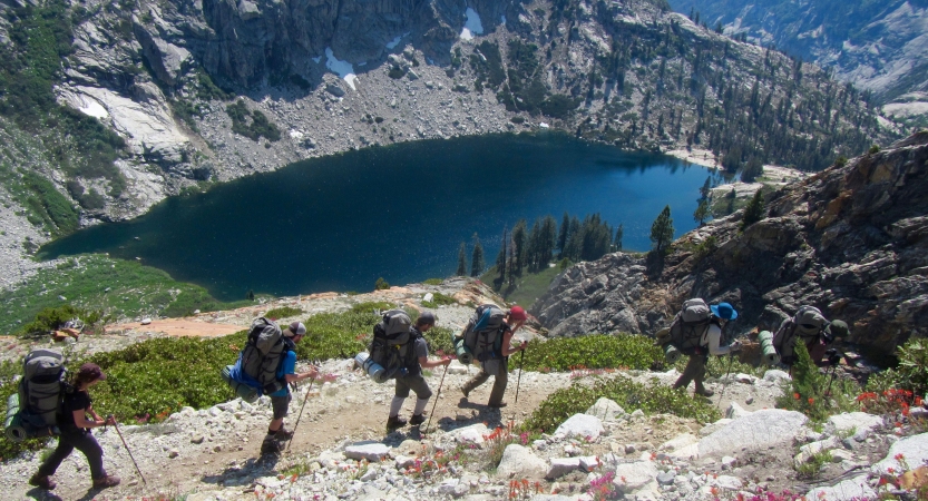 A group of people wearing backpacks hike near an alpine lake, framed by mountains. 
